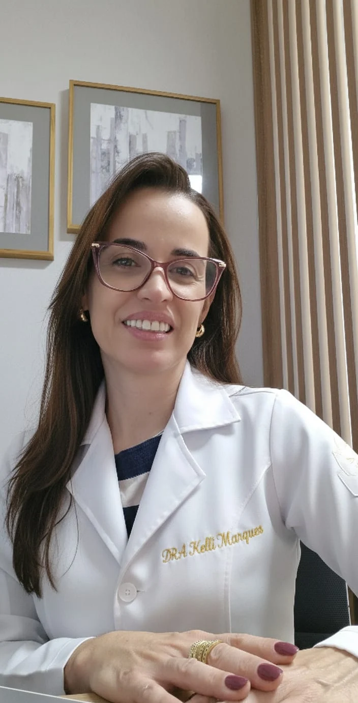 Dra. Kelli Marques à mesa do consultório, de óculos e jaleco com o nome bordado em dourado, sorrindo